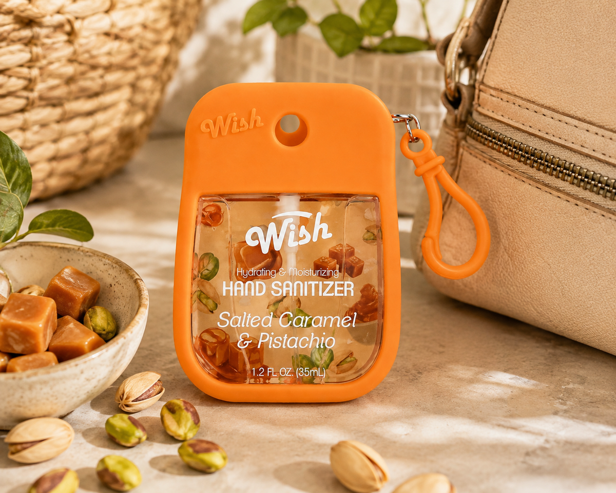 An orange Wish brand hand sanitizer bottle with a clear front panel showing salted caramel and pistachio pieces, styled on a marble surface with a woven basket, nuts, and a beige handbag nearby.