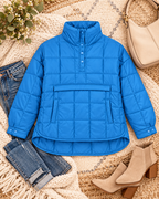 Flat lay of blue quilted puffer pullover jacket styled with jeans, tan ankle boots, and a cream bag on a woven blanket
