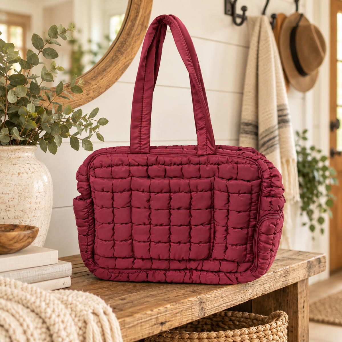 Wine Quilted Puffer Tote Bag on rustic wood entryway console table showing rich wine burgundy grid-quilted puffer nylon and dual flat padded handles beside speckled white ceramic vase with eucalyptus, wooden bowl, stacked books, and round gold mirror against white shiplap wall with farmhouse coat hooks, cream linen throw, and tan hat
