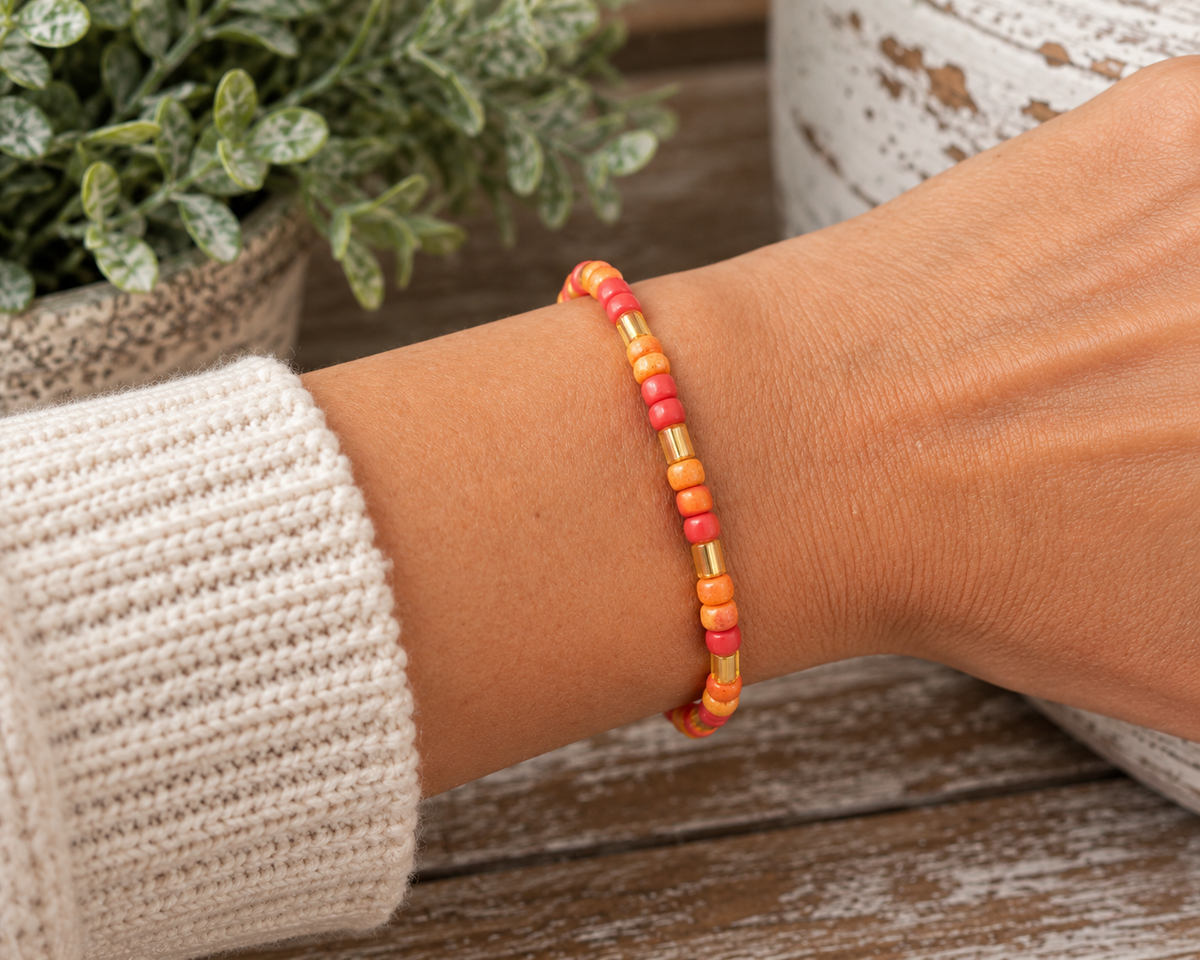 Woman wearing coral and orange seed bead stretch bracelet on wrist with cream crochet knit sleeve, concrete pot with greenery, and chippy white pedestal backdrop

