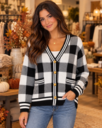 Brunette woman smiling in black and white plaid knit cardigan with antique gold dome buttons and striped trim, layered over white tank with blue jeans, in boutique interior with fall floral arrangements and pumpkins
