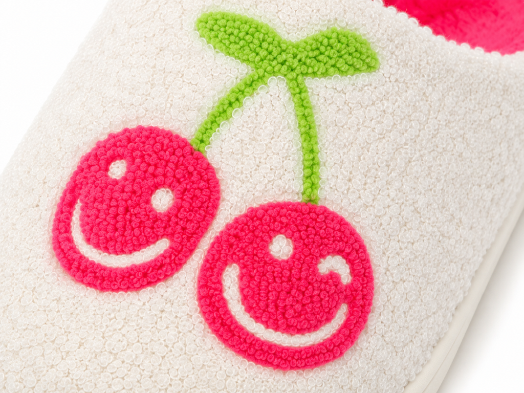 Close-up of pink smiley face cherry tufted design with green stem on white cozy slipper
