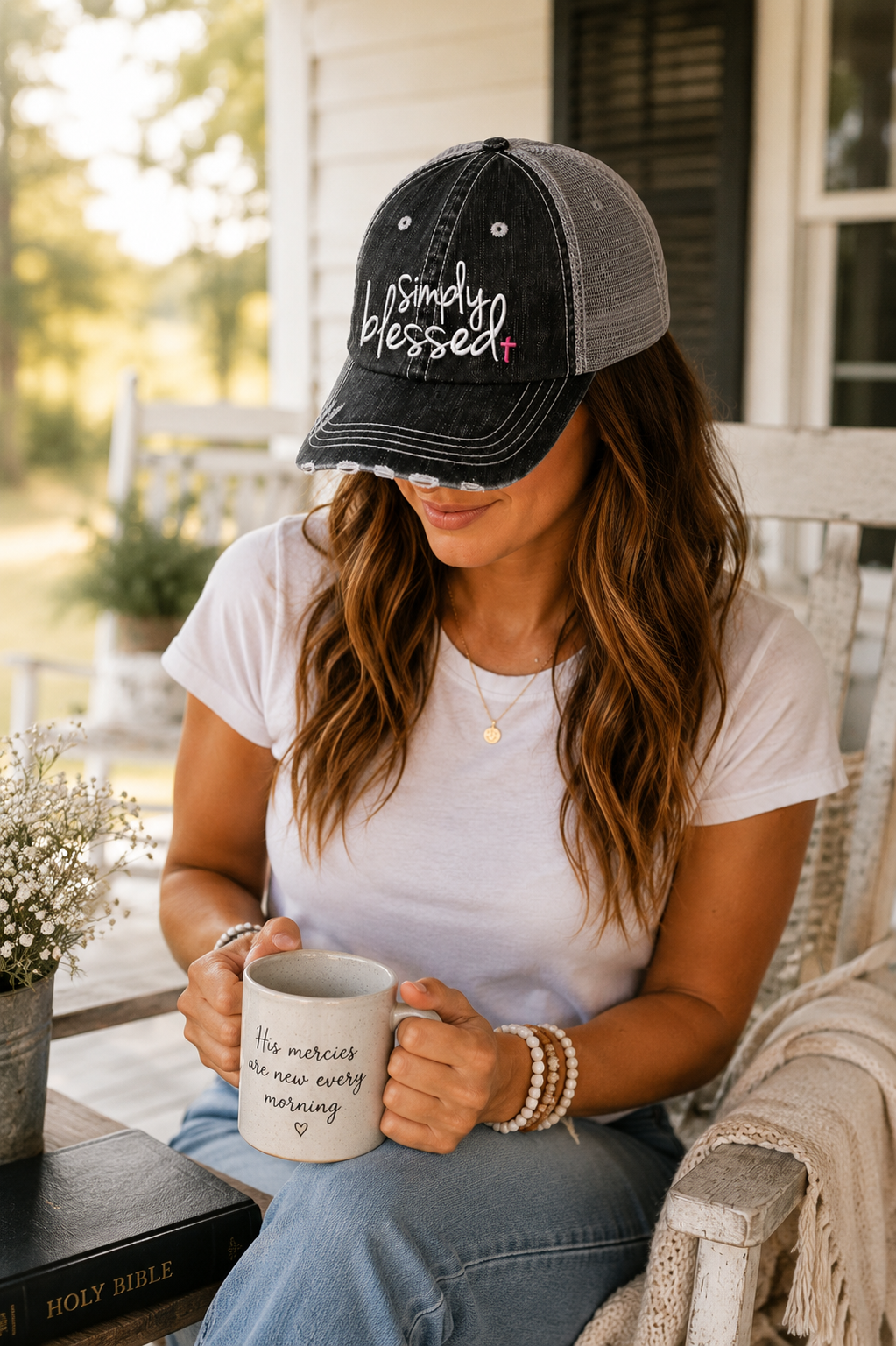 A woman sitting on a porch wearing a black and gray trucker hat with 'Simply Blessed' embroidered on the front, holding a white mug with black text.