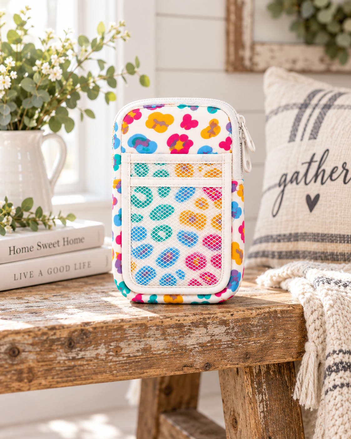 Multicolor Floral Tumbler Pouch on rustic wood bench showing white all-over multicolor retro floral print, white side zipper, front slip pocket, and white mesh pocket beside white pitcher of white flowers, stacked Home Sweet Home and Live a Good Life books, and grain sack gather pillow
