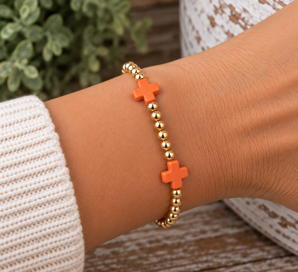 Woman wearing gold beaded stretch bracelet with orange cross accents on wrist with white lace knit sleeve, rustic wood surface, and greenery backdrop
