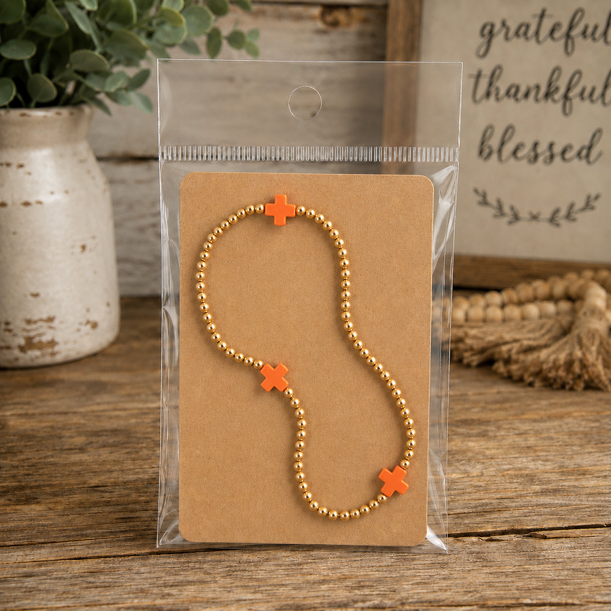 Gold beaded bracelet with orange cross accents on kraft card in clear packaging on rustic wood surface with chippy white vase, greenery, and grateful thankful blessed wood sign
