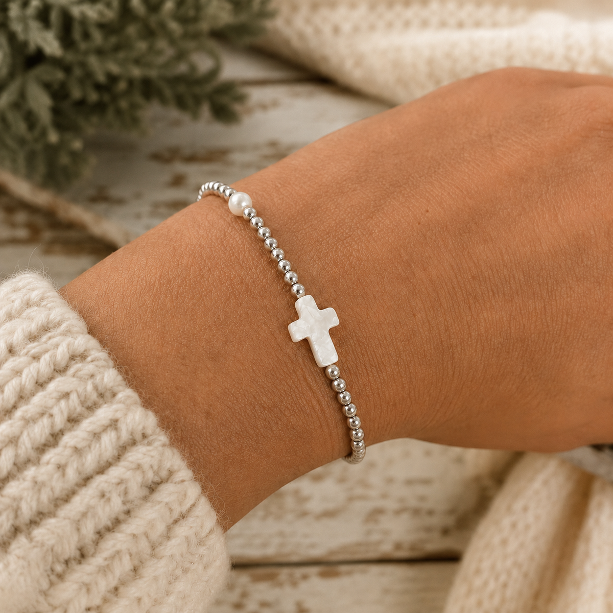 Woman wearing silver beaded cross bracelet with pearl accents on wrist with cream knit sweater and rustic chippy white wood backdrop
