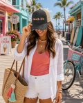 A woman stands on a sunny boardwalk wearing a black and beige trucker hat with 'beach life' embroidered in colorful letters. She is dressed in a white button-up shirt, a coral pink ribbed tank top, and white denim shorts, carrying a large straw tote bag.