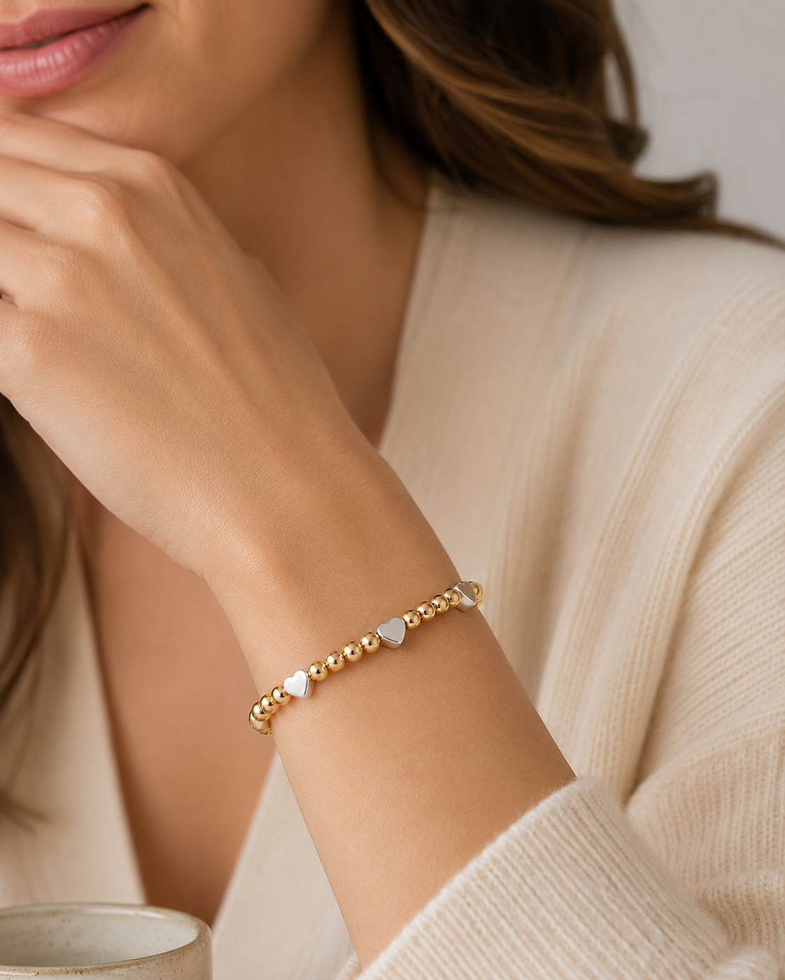 Woman wearing gold and silver heart beaded stretch bracelet on wrist with neutral cream sweater

