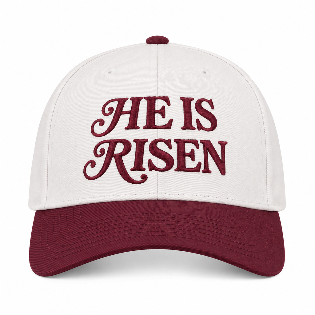 He Is Risen Baseball Cap in Raisin front view showing cream cotton crown with deep burgundy decorative script He Is Risen embroidery, solid raisin bill, and raisin top button on white background
