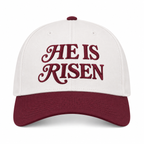He Is Risen Baseball Cap in Raisin front view showing cream cotton crown with deep burgundy decorative script He Is Risen embroidery, solid raisin bill, and raisin top button on white background
