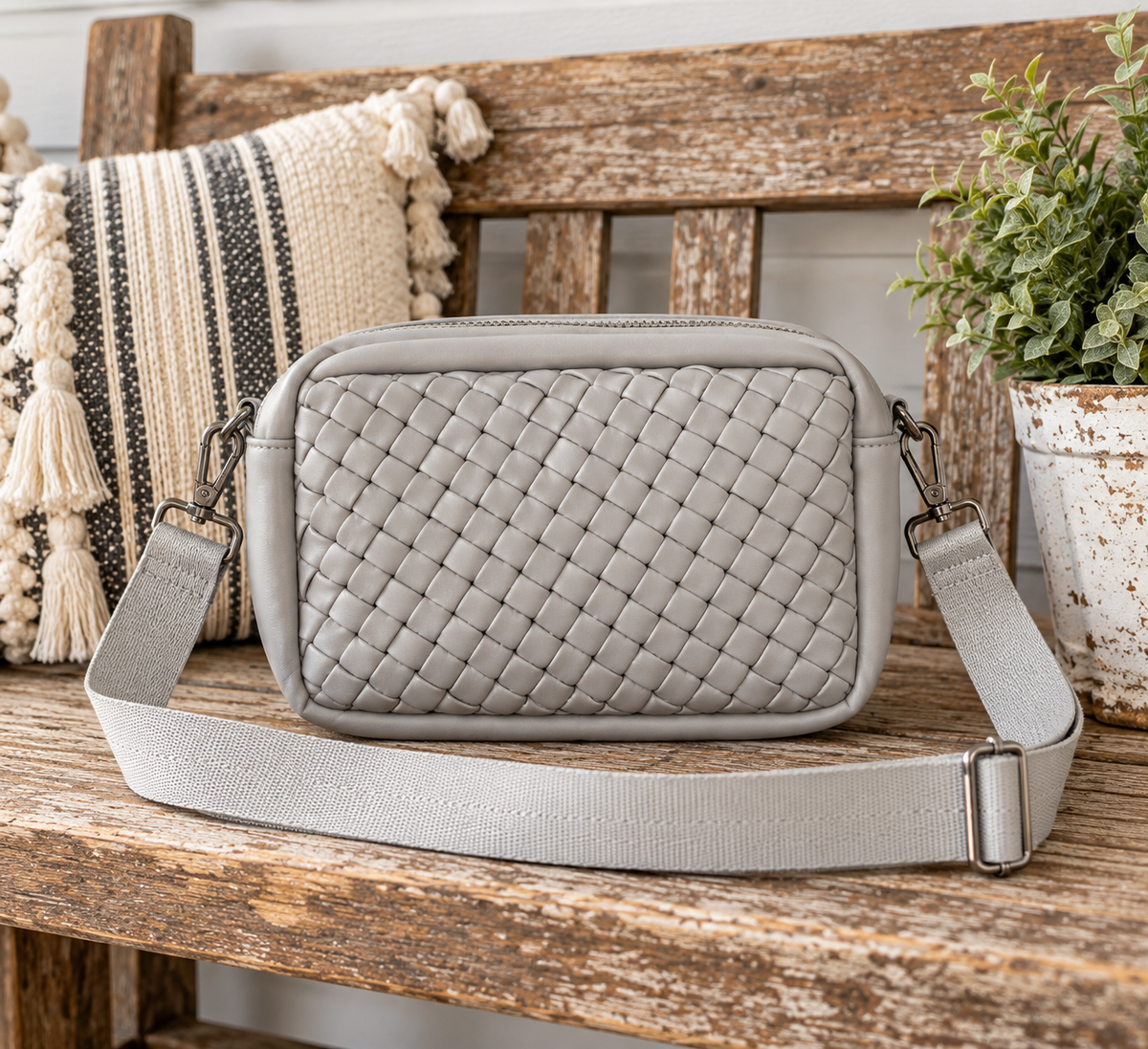 Gray Woven Crossbody Bag displayed on rustic wooden bench showing light gray interlaced woven panel, smooth gray trim, and adjustable wide gray webbing strap beside striped tasseled pillow and white distressed clay pot with greenery in farmhouse setting
