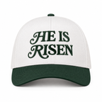 He Is Risen Baseball Cap in Dark Forest front view showing cream cotton crown with dark forest green decorative script He Is Risen embroidery, solid dark green bill, and dark green top button on white background
