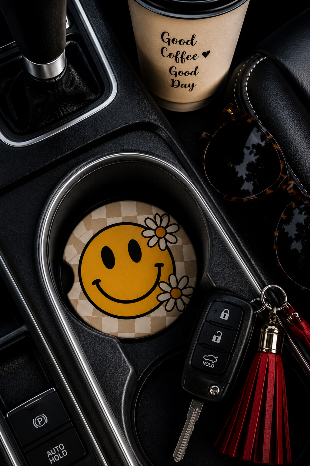 Smiley Face Daisy Car Coaster sitting in a car cup holder next to a coffee cup, sunglasses, and keys, showing the yellow smiley face and daisy design on a tan checkered background.
