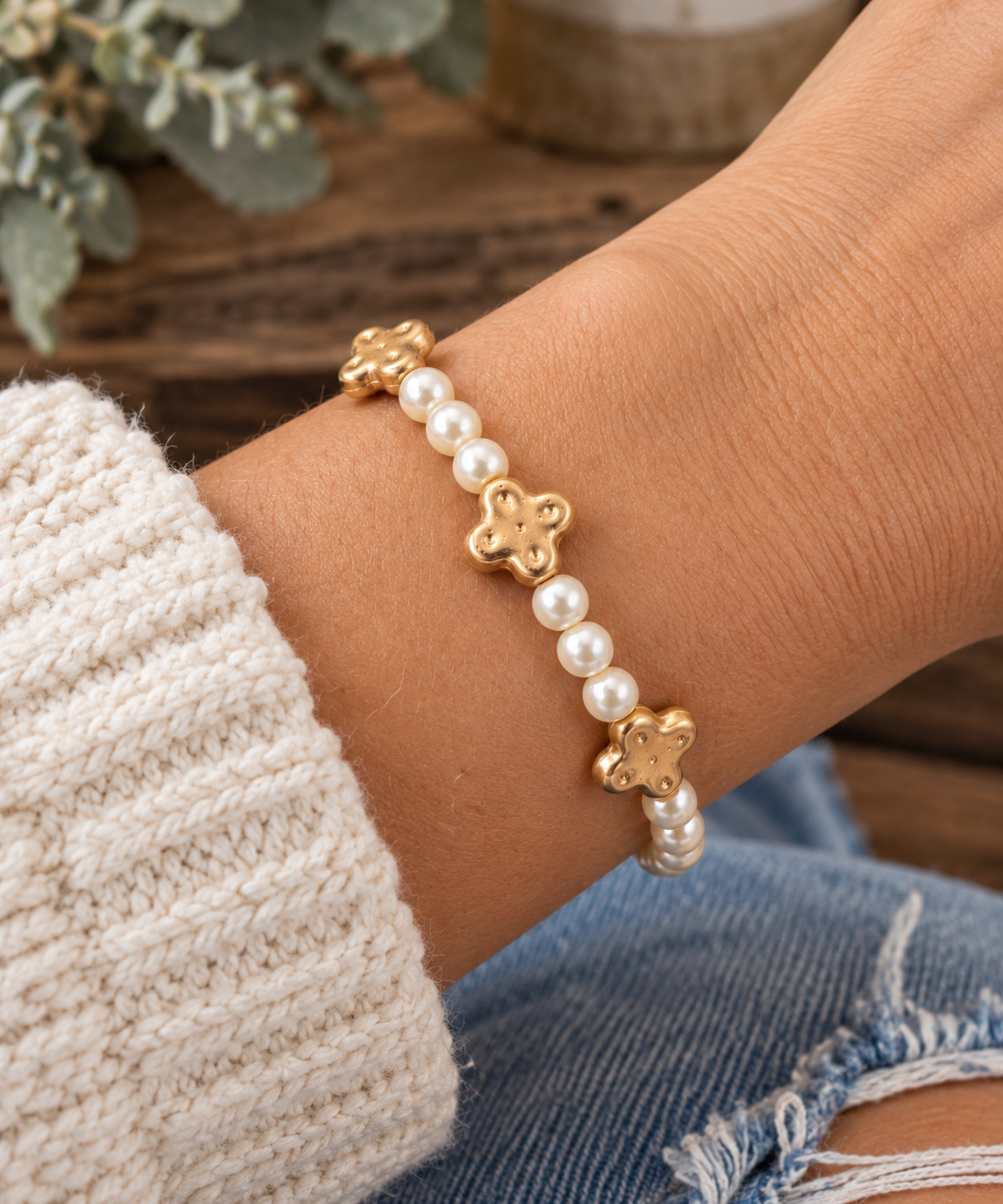 Woman wearing pearl beaded stretch bracelet with gold clover accents on wrist with cream knit sweater, distressed denim, and rustic wood and eucalyptus backdrop
