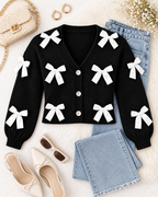 Black Satin Bow Knit Cardigan styled in flat lay on white textured surface with blue jeans, cream bow ballet flats, cream woven chain-strap crossbody bag, gold hoop earrings, brown square sunglasses, and baby's breath florals
