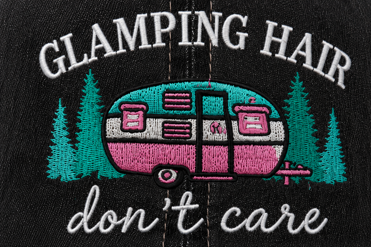 Close-up of embroidered "Glamping Hair Don't Care" lettering with colorful camper scene on a distressed black trucker hat front panel.
