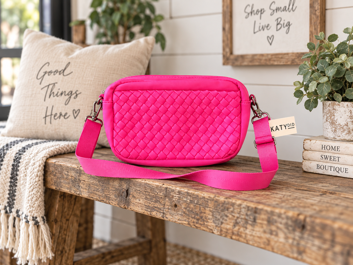 Hot Pink Woven Crossbody Bag displayed on rustic wooden bench showing vivid hot pink quilted diamond woven panel and adjustable wide hot pink strap with Katydid hang tag beside linen pillow, stacked books, and potted greenery in farmhouse boutique setting

