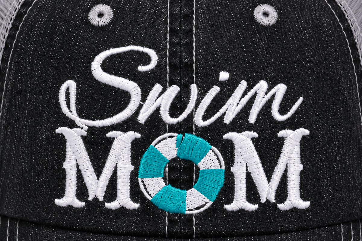 Close-up of white embroidered "Swim MOM" script with teal life preserver accent on a distressed black trucker hat front panel.
