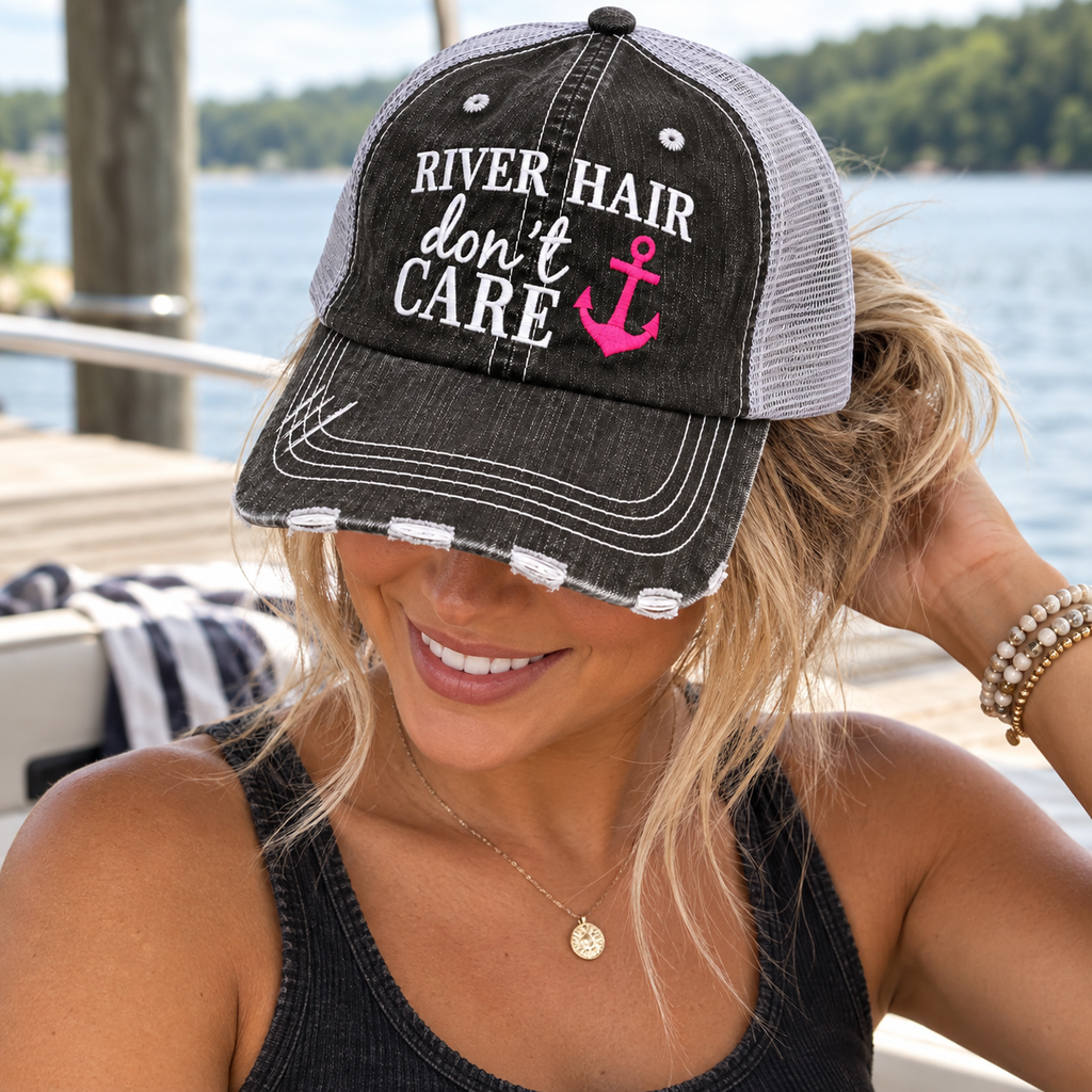 Woman wearing River Hair Don't Care Trucker Hat with distressed black front and gray mesh back showing white and pink embroidered River Hair Don't Care lettering with hot pink anchor with black tank and waterfront dock backdrop
