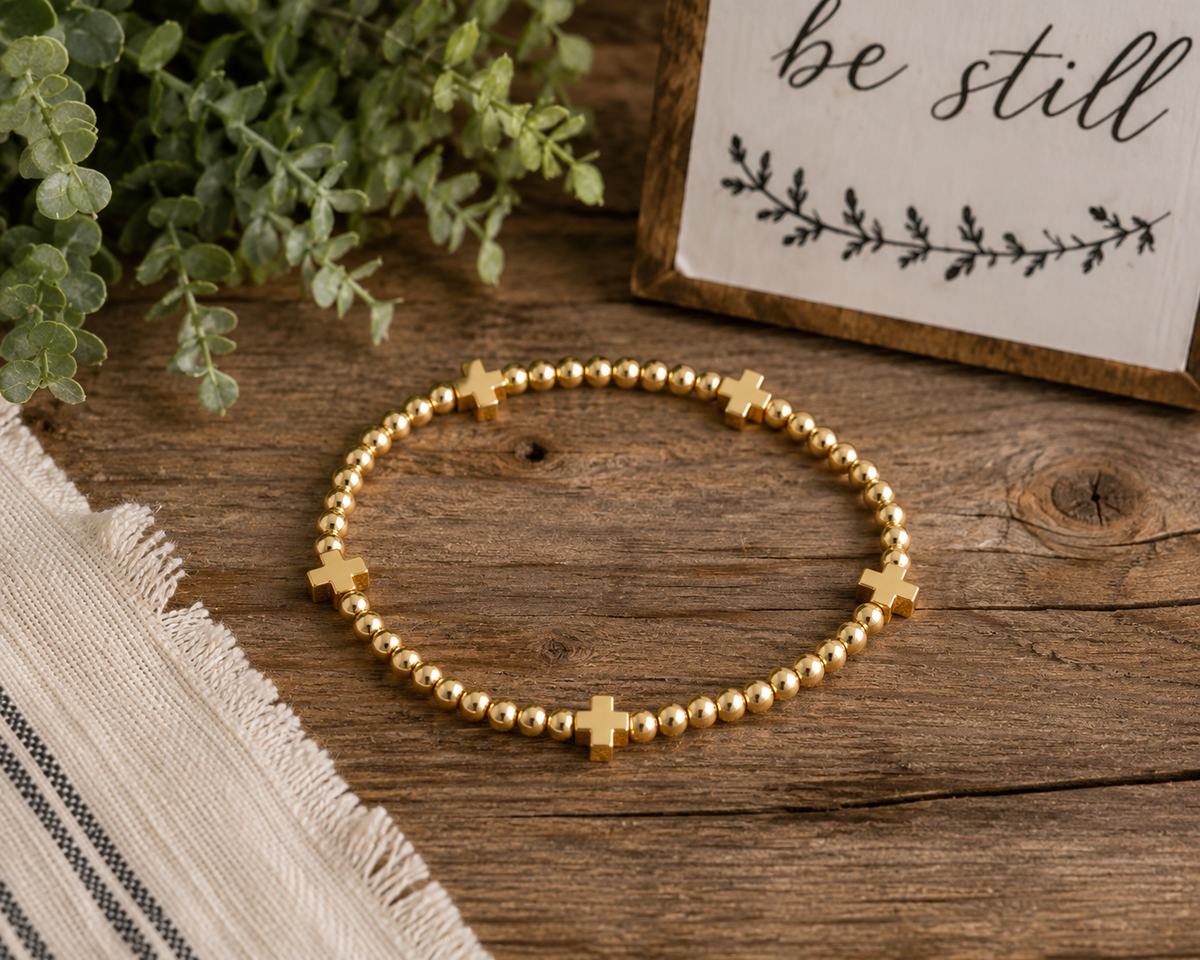 Gold cross beaded stretch bracelet laid flat on rustic wood surface with potted greenery, linen textile, and be still sign backdrop
