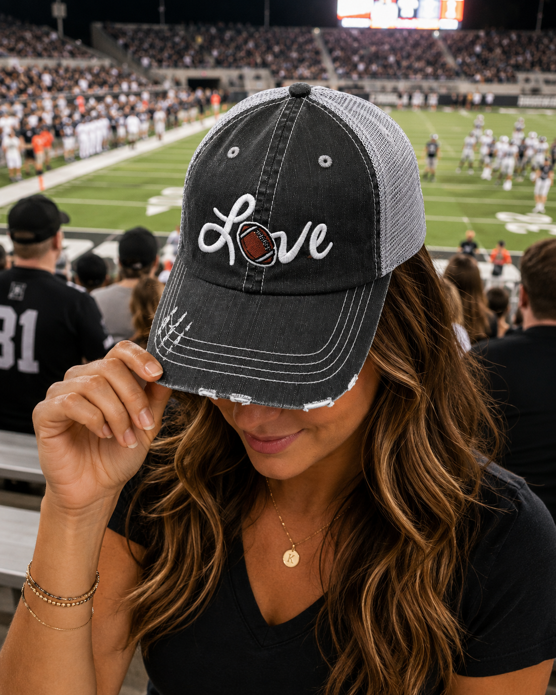 A woman in a stadium wearing a distressed charcoal and gray trucker hat with 'Love Football' embroidered on the front, featuring a football replacing the letter O, holding the brim up over her eye.