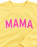 Close-up of pink MAMA lettering with a white outline on the yellow sweatshirt, showing the ribbed neckline and cuff
