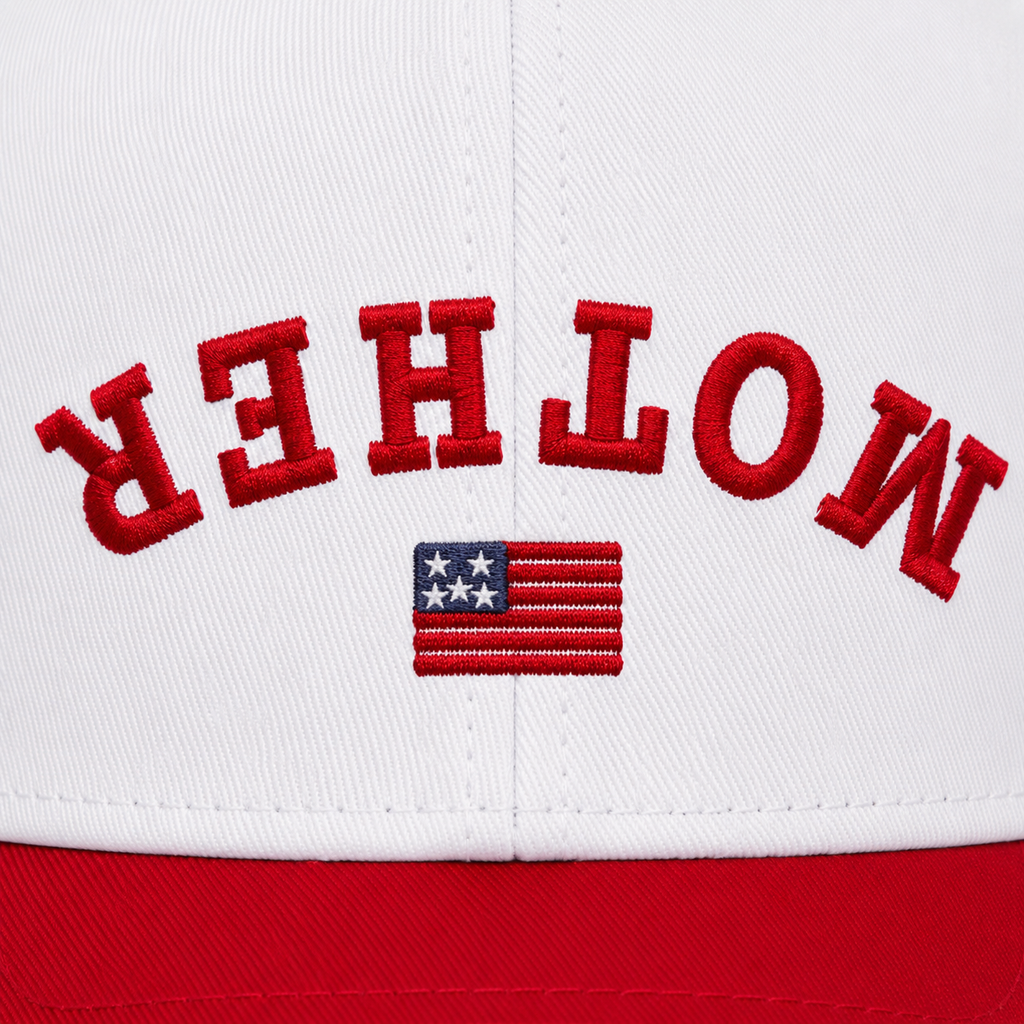 Close-up of bold red collegiate arched "MOTHER" embroidery with American flag accent below, intentionally flipped to read correctly when the brim is flipped up, on a white baseball cap front panel.
