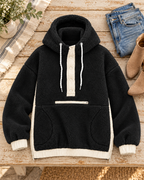Black two-toned sherpa pullover hoodie laid flat on a wooden table beside jeans and tan ankle boots, showing ivory trim and front pockets
