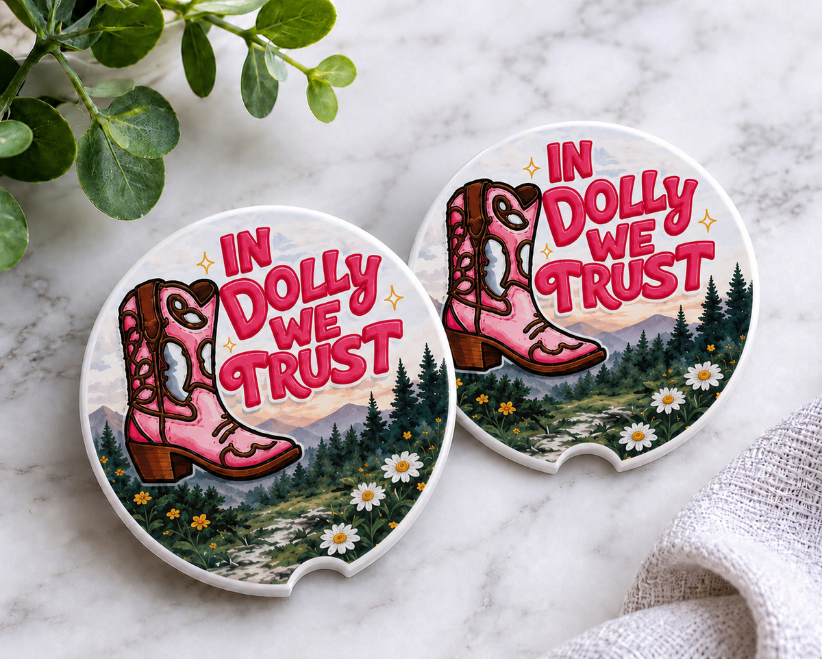 In Dolly We Trust Car Coaster Set — set of 2 ceramic coasters with pink cowgirl boot, mountain meadow, wildflowers, and "In Dolly We Trust" text in bold pink lettering, on marble surface
