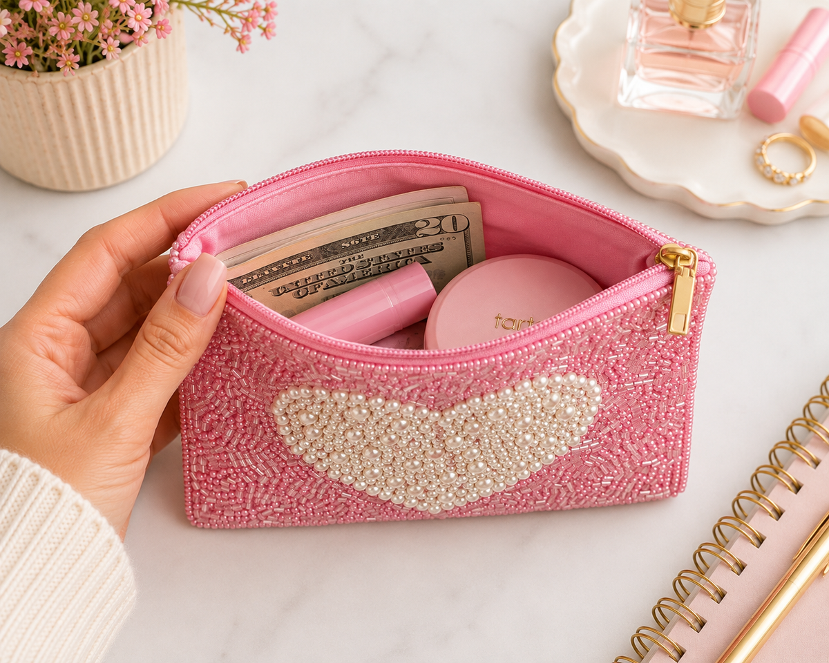 A pink beaded cosmetic pouch with a pearl-embellished heart on the front, held open by a hand. The pouch contains cash, a pink item, and a round compact.