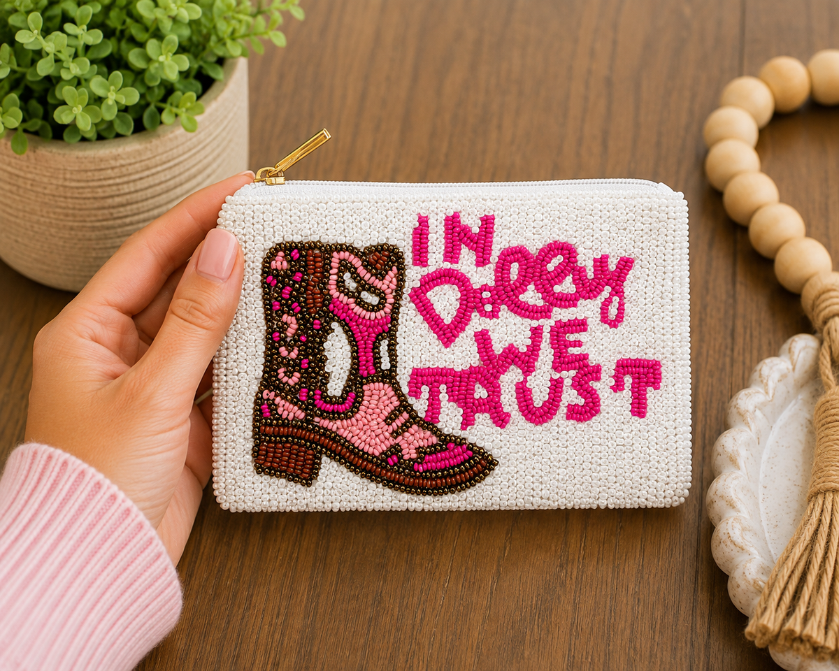 A hand holds a small white woven coin purse with a gold zipper. The purse features a brown and pink cowboy boot design and the phrase 'IN DOLLY WE TRUST' in pink lettering.