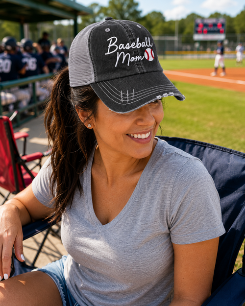 A woman sitting in a baseball stadium wearing a distressed black and gray trucker hat with 'Baseball Mom' embroidered on the front, paired with a gray v-neck t-shirt and denim shorts.