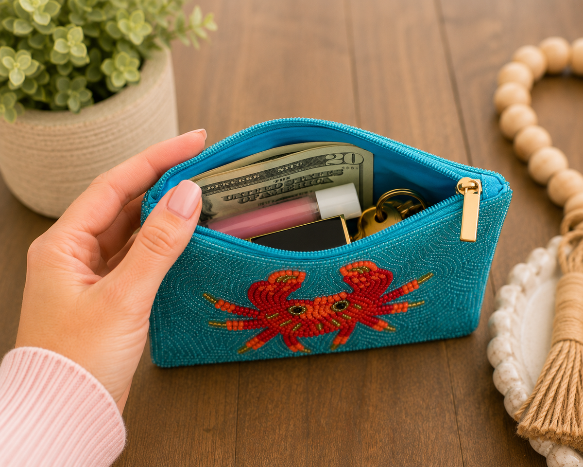 A turquoise blue woven zip pouch with a red and orange embroidered crab design, held open by a hand on a wooden tabletop. The pouch contains cash, a lip balm tube, and keys, with a potted plant and decorative beads in the background.
