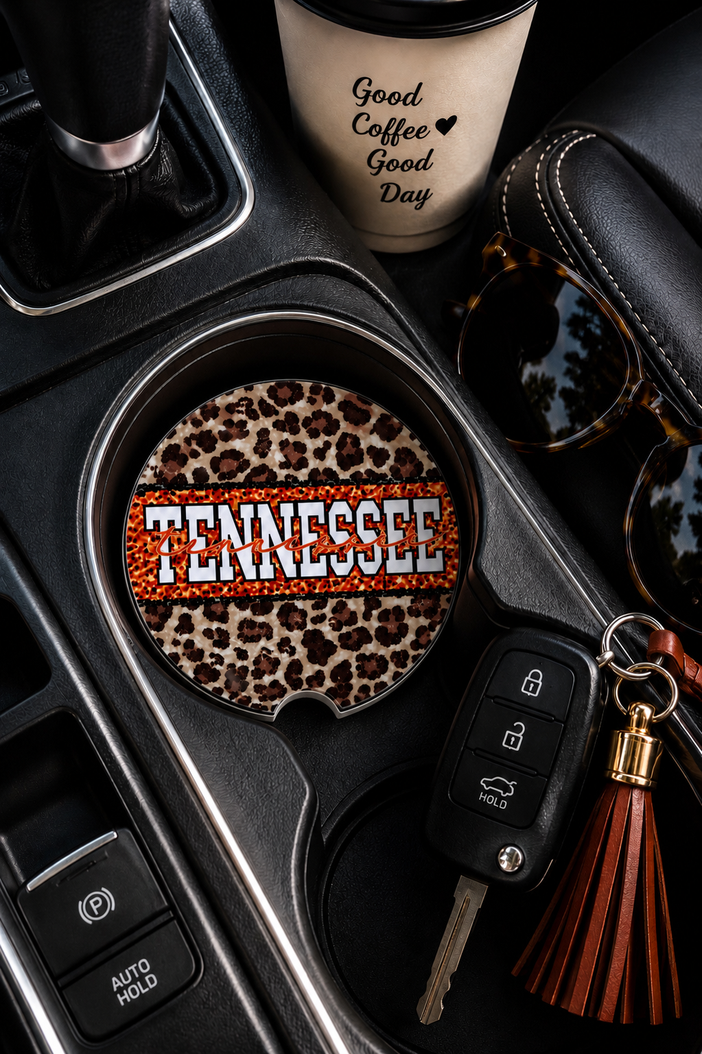 Tennessee Leopard ceramic car coaster sitting in vehicle cup holder next to a coffee cup, sunglasses, and keys — leopard print with bold orange Tennessee lettering
