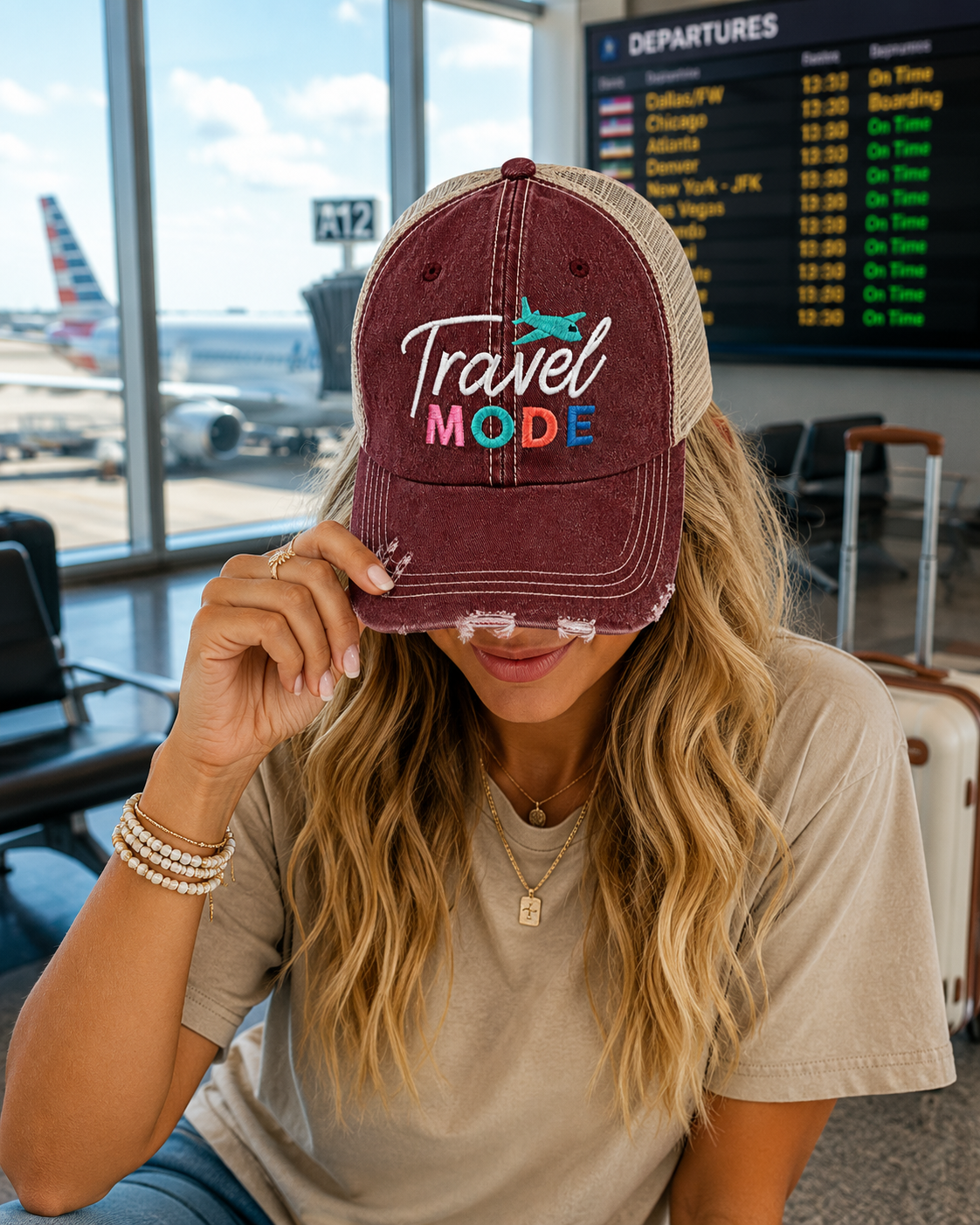 A woman in an airport terminal wearing a maroon and beige trucker hat with 'Travel Mode' embroidered on the front, holding the brim over her eyes.