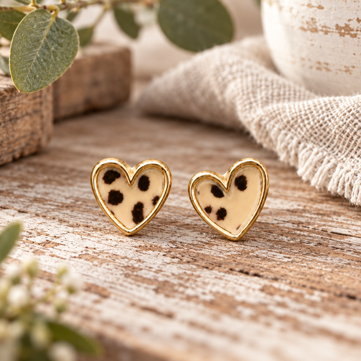 Leopard heart stud earrings with gold-tone trim displayed on whitewashed wood surface with eucalyptus, chippy white vase, and cream burlap fringe textile
