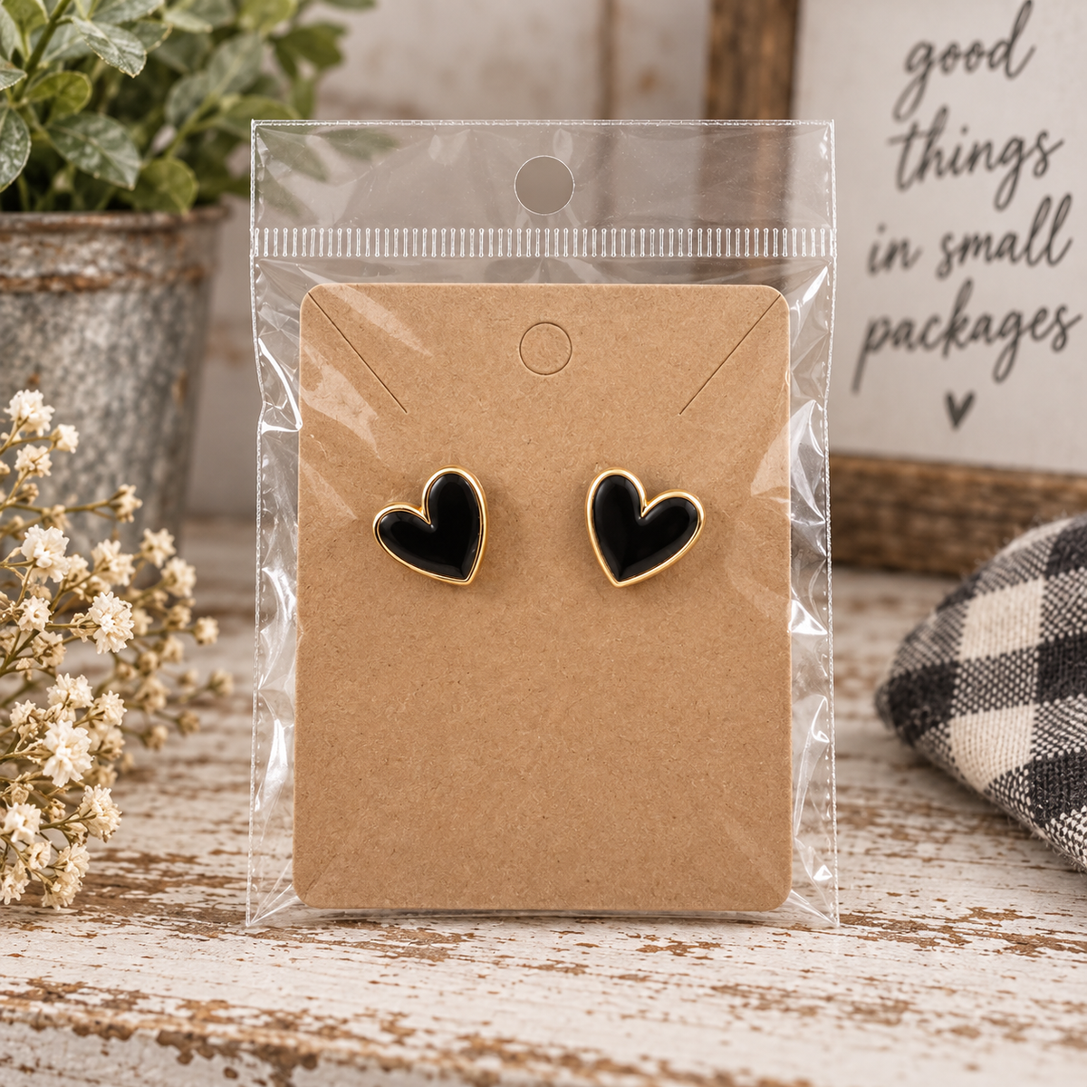 Black heart stud earrings with gold-tone trim on kraft card in clear packaging on chippy white wood with baby's breath, Good Things in Small Packages sign, buffalo check textile, and galvanized pot with greenery
