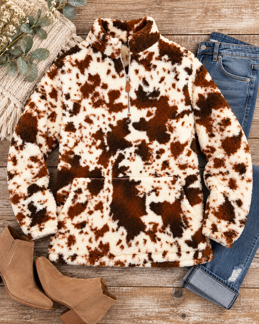 Brown and ivory cow print sherpa pullover styled with distressed jeans, tan ankle boots, a cream knit textile, and greenery on rustic wood
