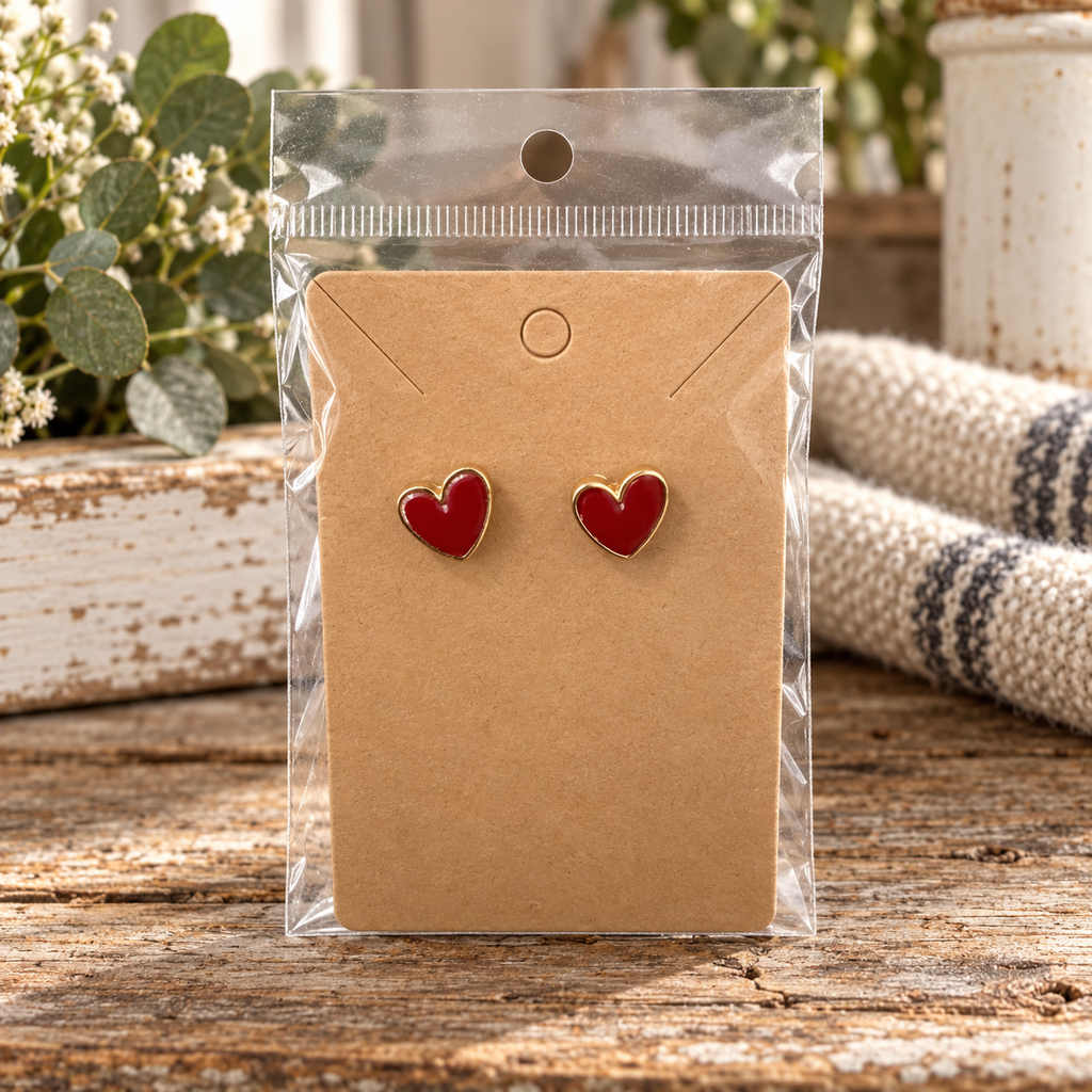 Red heart stud earrings with gold-tone trim on kraft card in clear packaging on rustic wood surface with eucalyptus and cream ticking stripe textile

