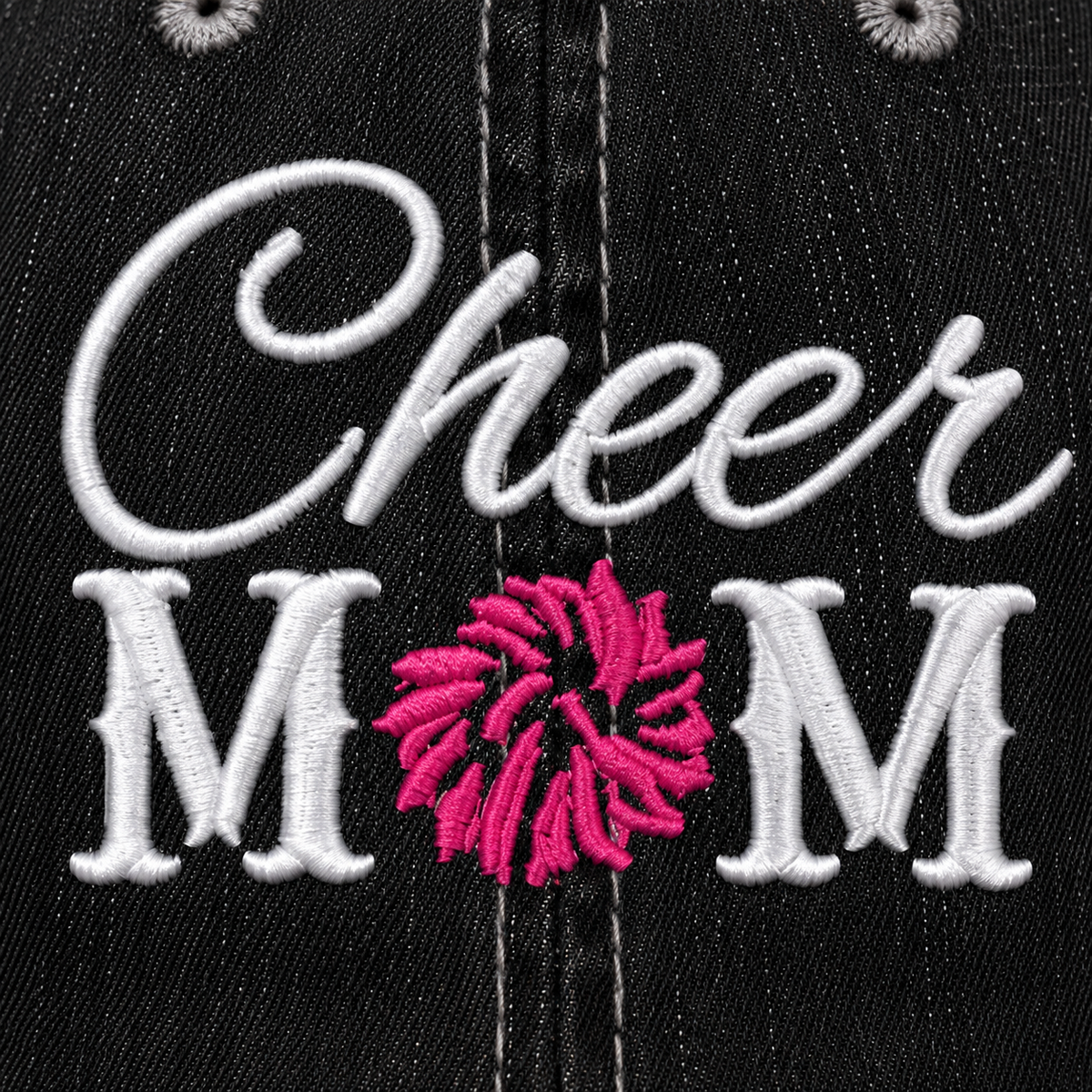 Close-up of white embroidered "Cheer MOM" lettering with pink pom-pom detail on a distressed black trucker hat front panel.
