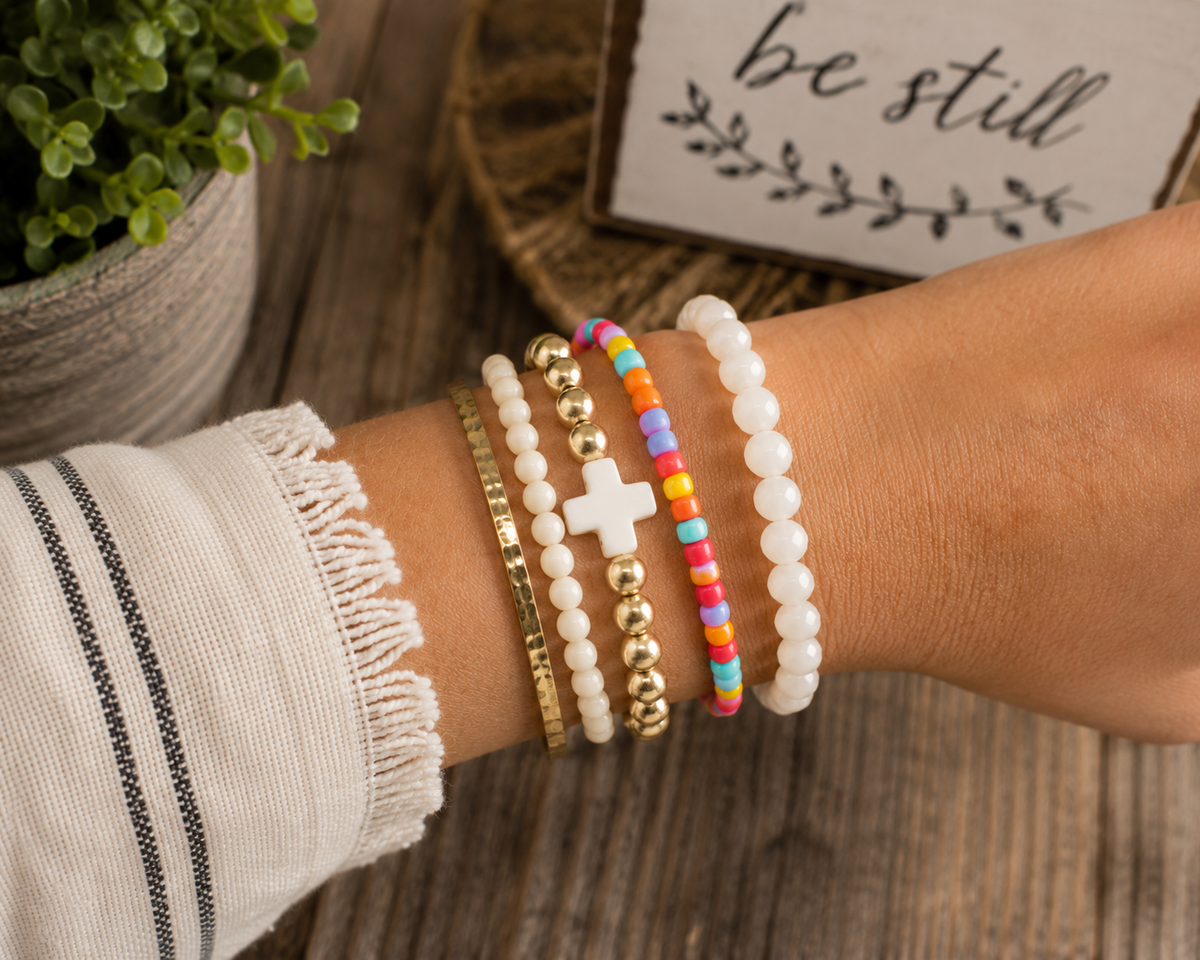 Colorful seed bead bracelet stacked with gold and white cross bracelet, white bead bracelet, and hammered gold bangle on wrist
