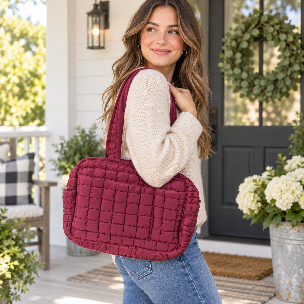 Woman in cream sweater and jeans carrying Wine Quilted Puffer Tote Bag on shoulder showing rich wine burgundy grid-quilted puffer nylon and dual flat padded handles on white farmhouse porch with black door, eucalyptus wreath, white hydrangeas, and buffalo check chair
