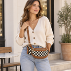 Brunette woman in cream V-neck ribbed cardigan and light wash jeans wearing Duo Checkered Crossbody Bag in Camel as crossbody showing black and cream checkered body with camel tan trim and gold zipper, outdoors near white stucco wall with olive tree and patio setting in background
