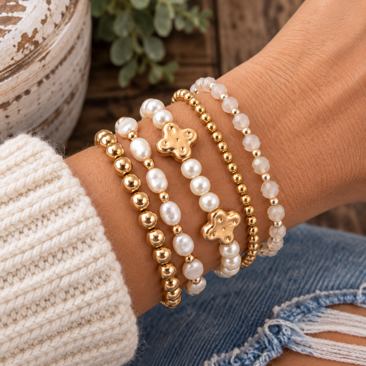 Pearl and gold clover bracelet stacked with pearl and gold ball bracelet, freshwater oval pearl bracelet, gold ball bracelet, and white crystal gold bead bracelet on wrist with cream knit sweater and rustic wood backdrop
