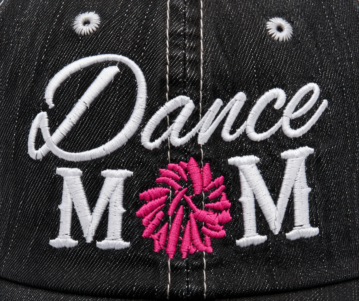 Close-up of white embroidered "Dance MOM" lettering with pink pom-pom detail on a distressed black trucker hat front panel.
