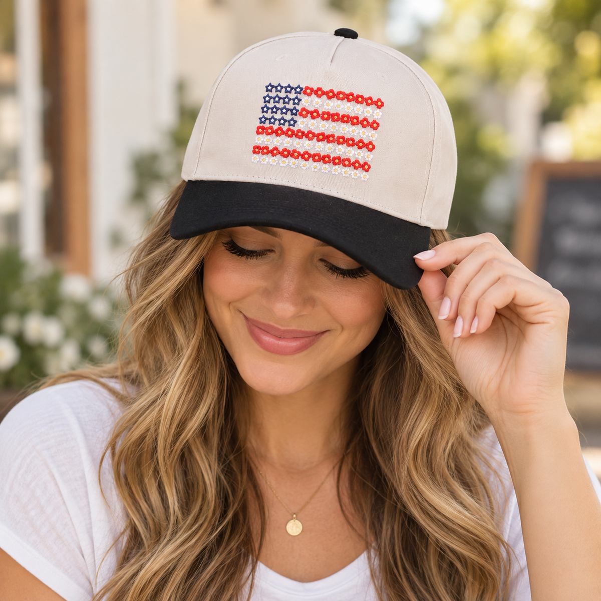 Brunette woman with wavy highlighted hair smiling down while tilting brim of Star Floral American Flag Baseball Cap in Black showing cream crown with embroidered floral flag and black bill, in boutique setting with chalkboard and brown felt hat in background
