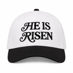 He Is Risen Baseball Cap in Black front view showing white cotton crown with black decorative script He Is Risen embroidery, solid black bill, and black top button on white background
