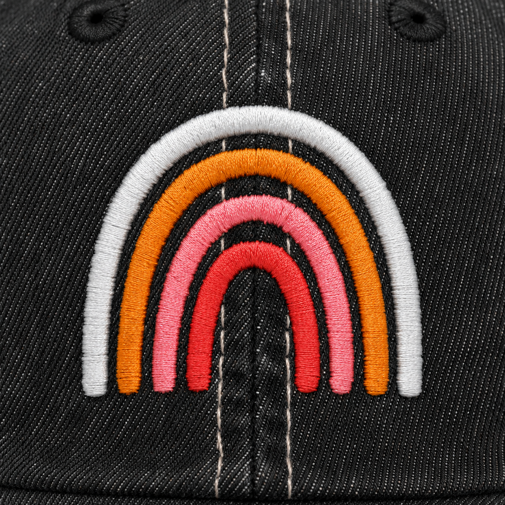 Close-up of embroidered boho rainbow design in soft white, orange, pink, and coral tones on a distressed black trucker hat front panel.
