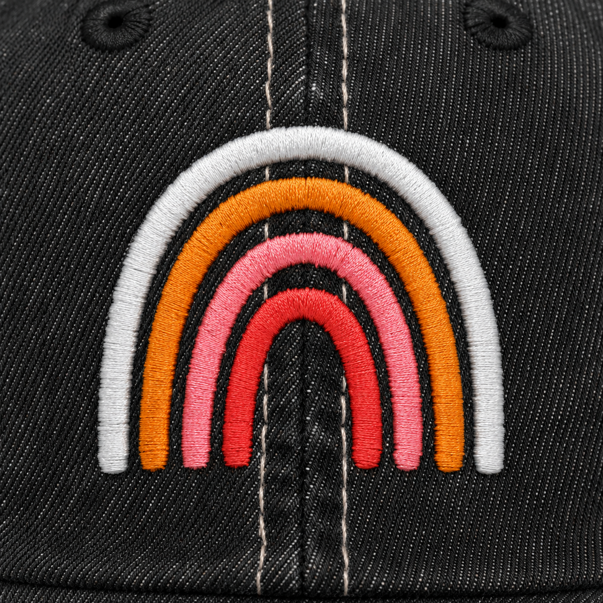 Close-up of embroidered boho rainbow design in soft white, orange, pink, and coral tones on a distressed black trucker hat front panel.
